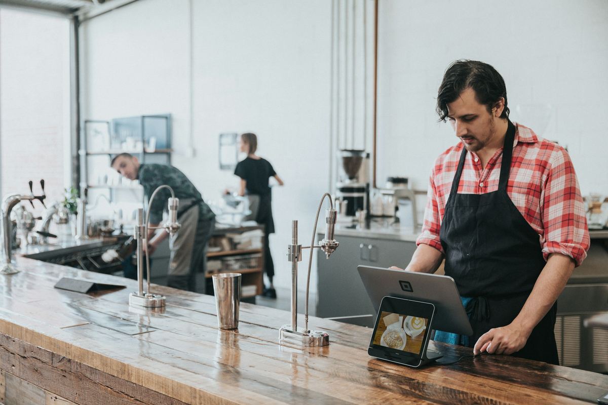A coffee shop owner in an apron checks the numbers on a tablet at the counter